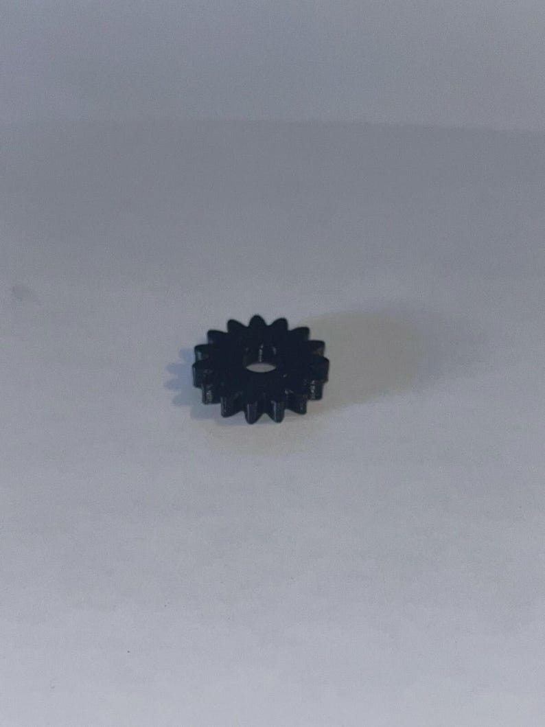 May include: A small, black, circular gear featuring twelve evenly spaced teeth around its perimeter and a single, centred circular hole. The component is constructed from a solid, dark material with a smooth finish and a uniform, symmetrical design.