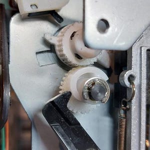 May include: Close-up of a white plastic gear mechanism with a black lever. The gears are interlocked and appear to be part of a larger machine.