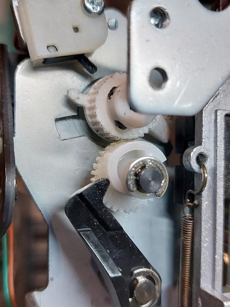 May include: Close-up of a white plastic gear mechanism with a black lever. The gears are interlocked and appear to be part of a larger machine.