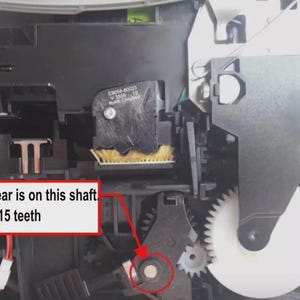 May include: Internal printer mechanism featuring a black plastic housing, a white circular gear, and a yellow brush component. A red arrow points to a gear on a shaft, accompanied by a red-bordered text box stating: The gear is on this shaft. It has 15 teeth.