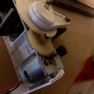 Może przedstawiać: Zbliżenie na biały silnik elektryczny z układem zębatym i czarnym zębatką. Silnik jest podłączony do czerwonych i czarnych przewod&oacute;w.