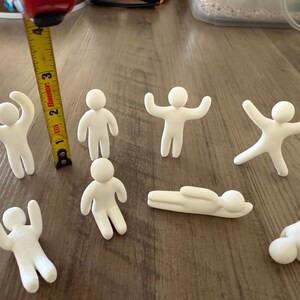 May include: Seven small white clay figures in various poses, including standing, sitting, and lying down. A yellow measuring tape is positioned next to one figure, showing a height of approximately 2.5 inches for the standing figures.