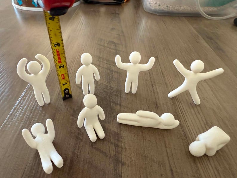 May include: Seven small white clay figures in various poses, including standing, sitting, and lying down. A yellow measuring tape is positioned next to one figure, showing a height of approximately 2.5 inches for the standing figures.