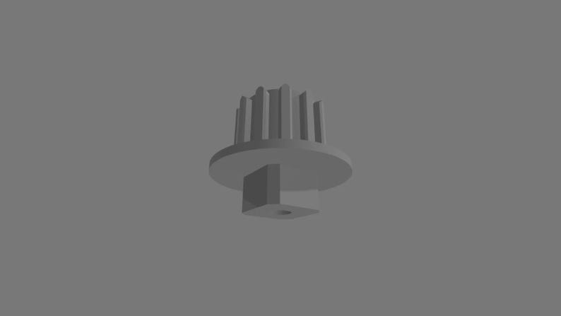 Puede incluir: Un componente de engranaje de pl&aacute;stico gris con una base circular, una serie de crestas verticales a lo largo del borde superior y una protuberancia cuadrada que se extiende desde el centro inferior, con un peque&ntilde;o orificio circular.