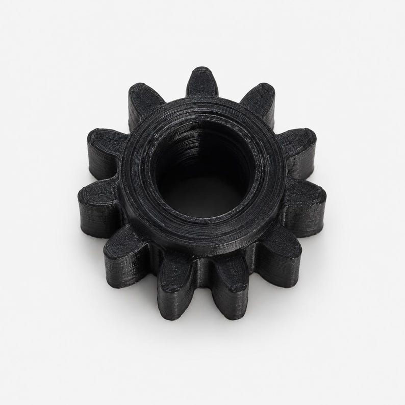 May include: A black gear with ten teeth and a circular center hole. The object features a textured surface with visible horizontal layers, indicating a 3D printed construction. The gear is circular with a uniform black finish throughout.