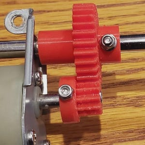 Op de afbeelding: Een mechanische assemblage met een rood kunststof tandwiel op een metalen as, verbonden met een kleiner rood tandwiel op een motorspil. De onderdelen zijn vastgezet met zilverkleurige zeskantbouten en bevestigd aan een metalen montagebeugel.