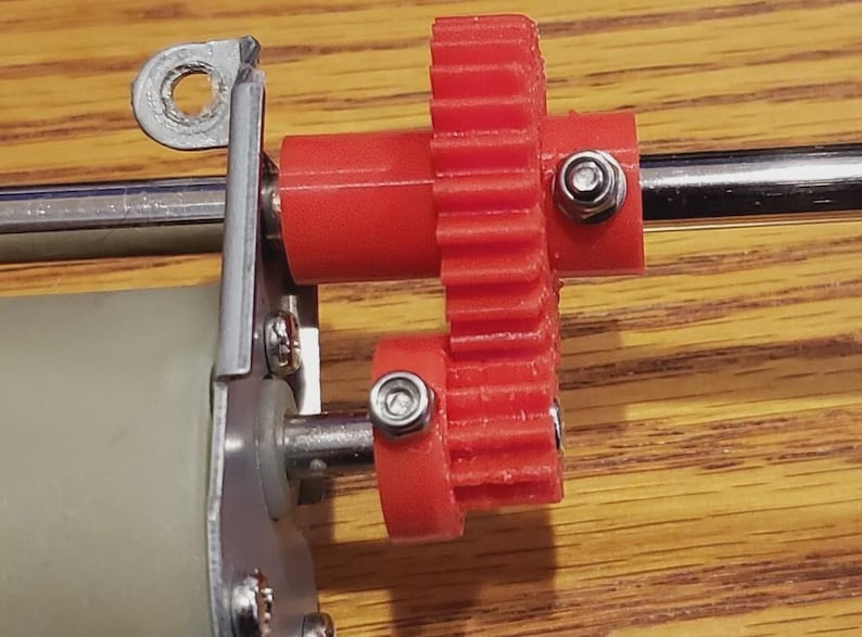 Op de afbeelding: Een mechanische assemblage met een rood kunststof tandwiel op een metalen as, verbonden met een kleiner rood tandwiel op een motorspil. De onderdelen zijn vastgezet met zilverkleurige zeskantbouten en bevestigd aan een metalen montagebeugel.