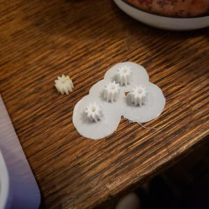 Może przedstawiać: Cztery białe plastikowe koła zębate na białym tle. Jedno mniejsze białe plastikowe koło zębate znajduje się na drewnianej powierzchni.