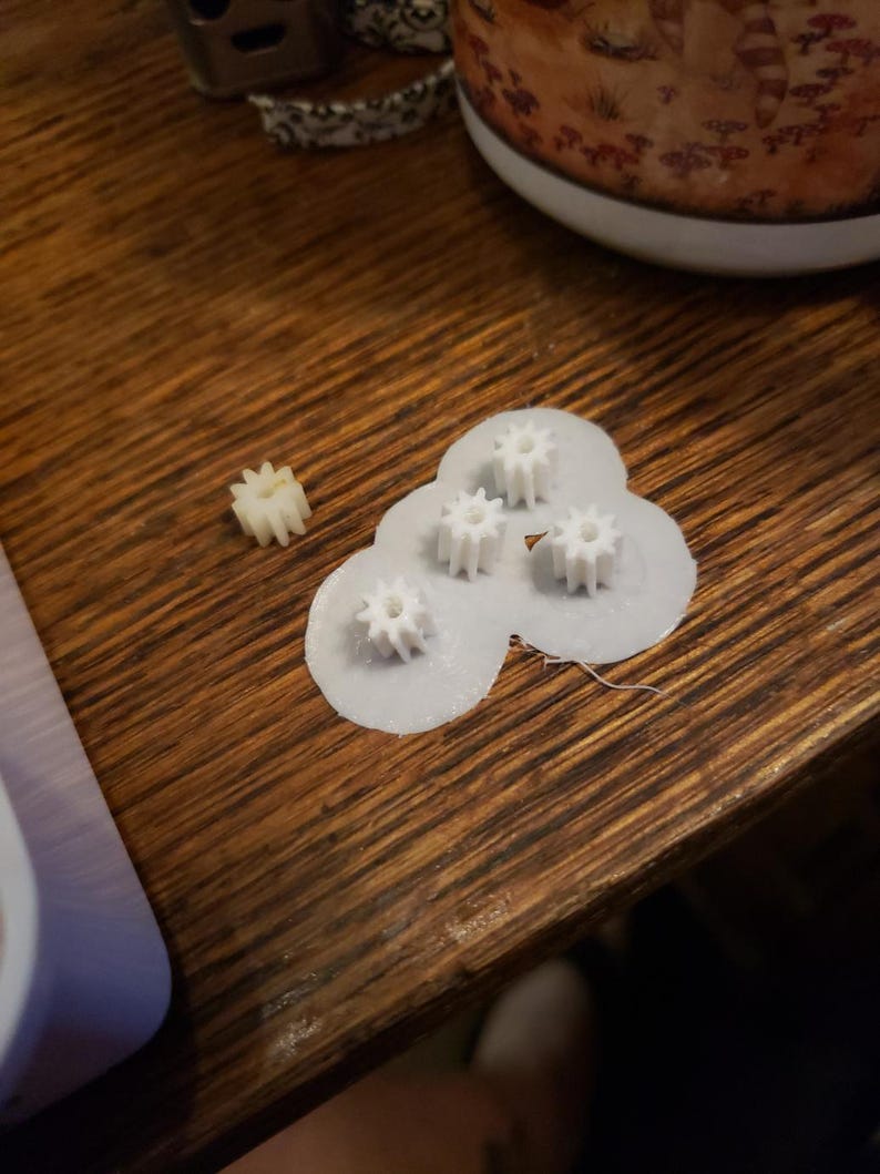 Może przedstawiać: Cztery białe plastikowe koła zębate na białym tle. Jedno mniejsze białe plastikowe koło zębate znajduje się na drewnianej powierzchni.