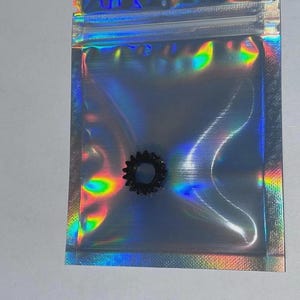 May include: A small, black, circular gear-shaped component with serrated edges, enclosed within a translucent, iridescent, holographic plastic zip-top storage bag that reflects a spectrum of rainbow colours across its surface.