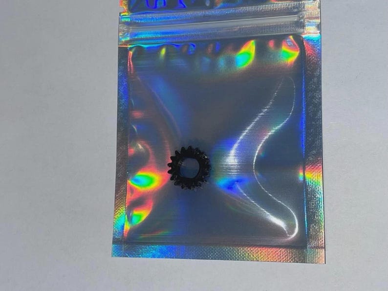 May include: A small, black, circular gear-shaped component with serrated edges, enclosed within a translucent, iridescent, holographic plastic zip-top storage bag that reflects a spectrum of rainbow colours across its surface.