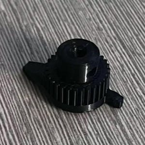 Puede incluir: Engranaje de plástico negro con un agujero central y un pequeño borde elevado alrededor del borde. El engranaje tiene una serie de dientes alrededor de su circunferencia.