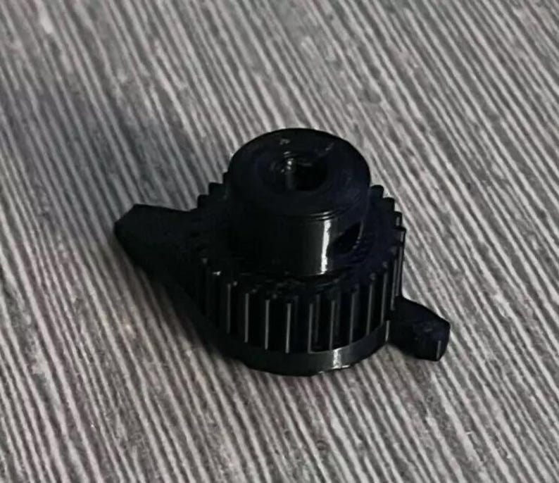 May include: Black plastic gear with a central hole and a small, raised lip around the edge. The gear has a series of teeth around its circumference.