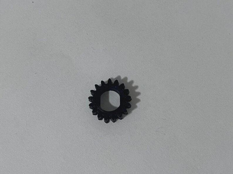 May include: A small, black, circular gear featuring a series of evenly spaced teeth around its outer edge and a central hole with a flat side, designed for mechanical rotation and power transmission within a larger assembly or device.