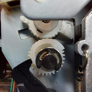 May include: Close-up of a white plastic gear mechanism with a black spring. The gears are interlocked and appear to be part of a larger machine.