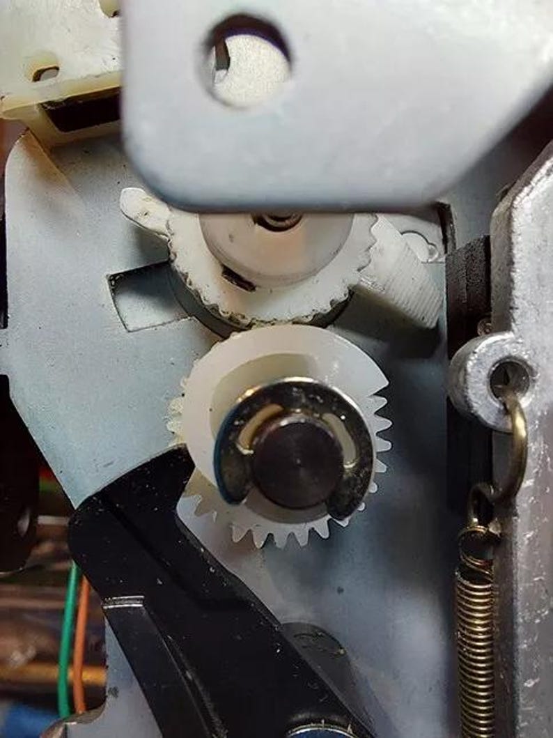 May include: Close-up of a white plastic gear mechanism with a black spring. The gears are interlocked and appear to be part of a larger machine.