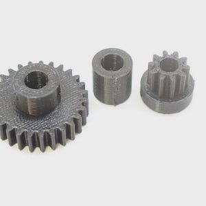 Op de afbeelding: Drie zwarte 3D-geprinte mechanische onderdelen bestaande uit een groot tandwiel met een centraal gat, een kleine cilindrische afstandhouder met een hol midden en een kleiner rondsel met een centraal gat en verticale tanden rondom.