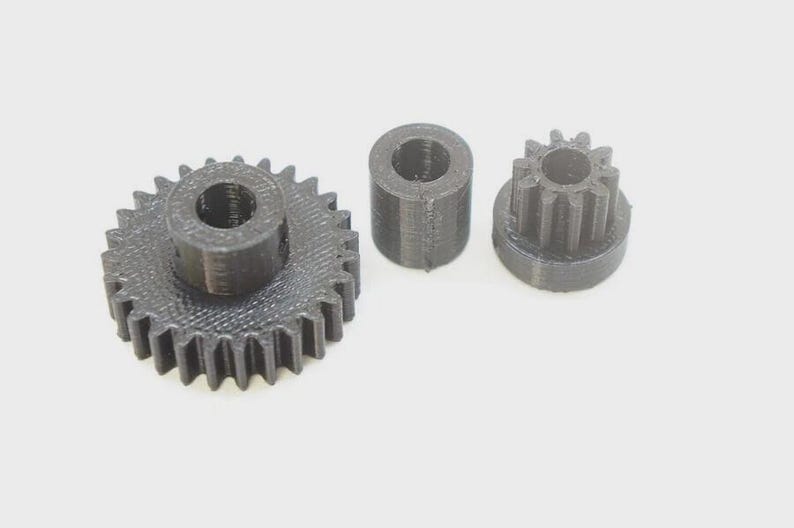 Op de afbeelding: Drie zwarte 3D-geprinte mechanische onderdelen bestaande uit een groot tandwiel met een centraal gat, een kleine cilindrische afstandhouder met een hol midden en een kleiner rondsel met een centraal gat en verticale tanden rondom.