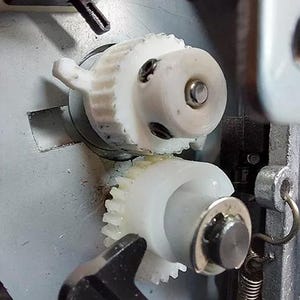 May include: Close-up of a white plastic gear mechanism with a metal spring. The gears are interlocked and appear to be part of a larger machine.