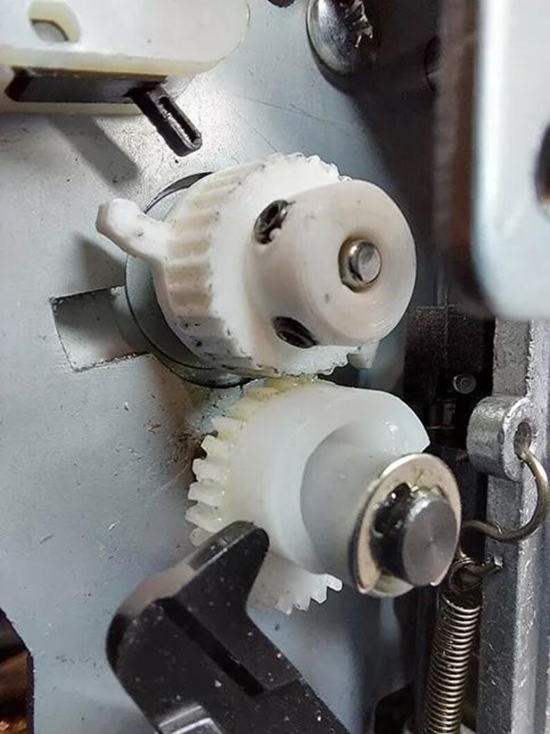 May include: Close-up of a white plastic gear mechanism with a metal spring. The gears are interlocked and appear to be part of a larger machine.