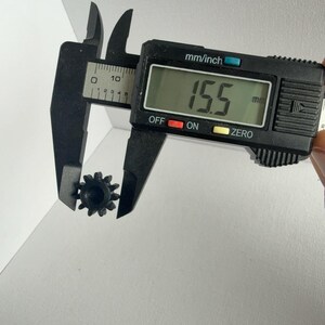 May include: A small black plastic gear with twelve teeth being measured by a black digital caliper. The digital display on the caliper clearly shows a measurement of 15.5 mm, indicating the outer diameter of the gear component.