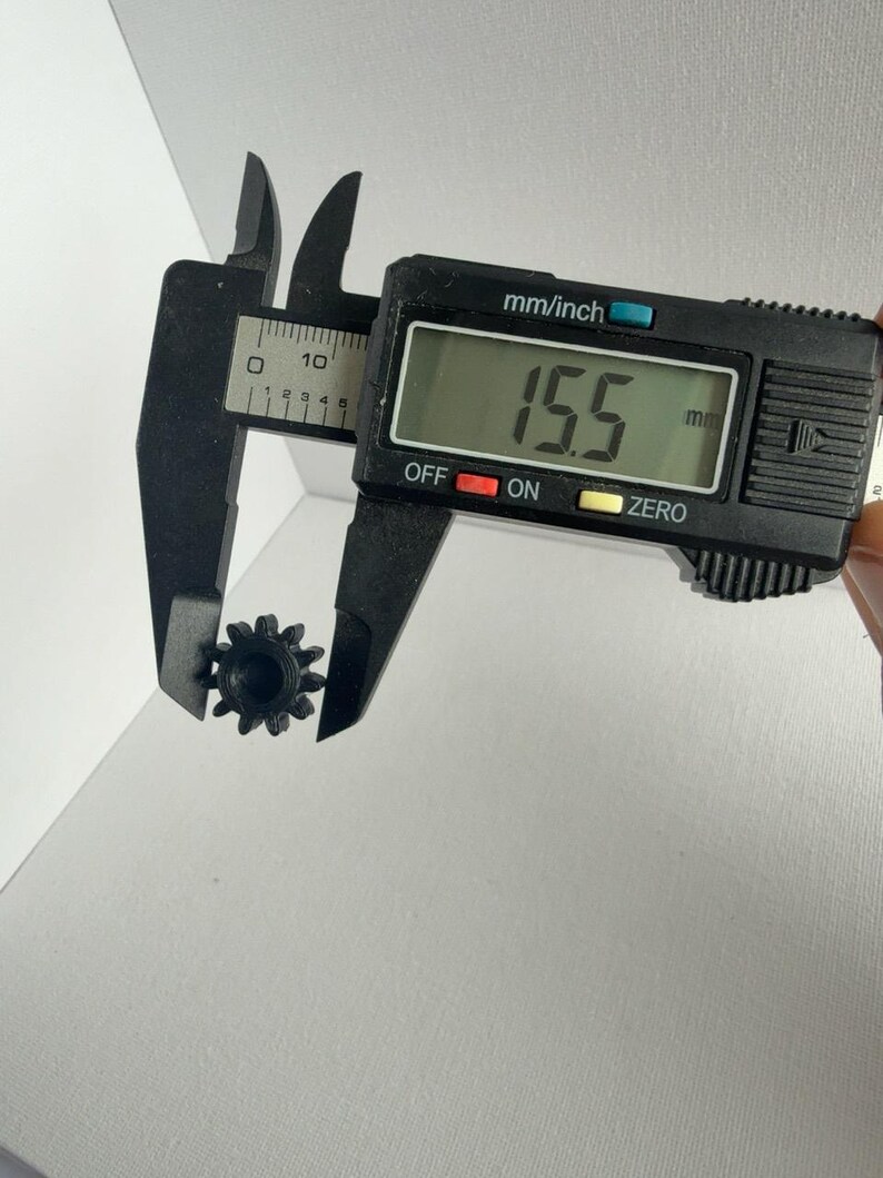 May include: A small black plastic gear with twelve teeth being measured by a black digital caliper. The digital display on the caliper clearly shows a measurement of 15.5 mm, indicating the outer diameter of the gear component.