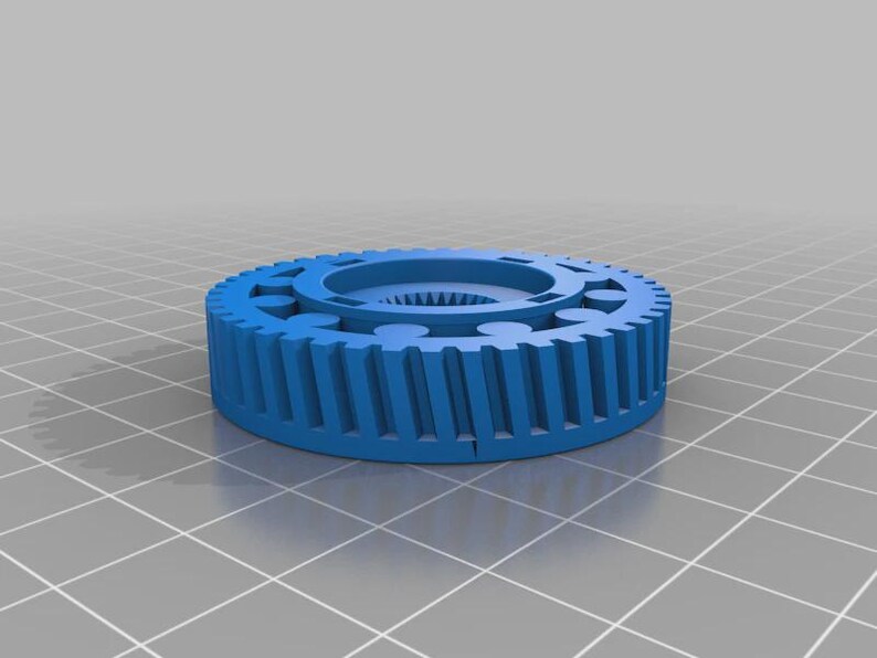 Op de afbeelding: Een blauw cirkelvormig tandwiel met een reeks verticale tanden langs de buitenrand en een centrale naaf met interne spiebanen. Het object heeft een plat bovenoppervlak met een ring van cirkelvormige inkepingen rond de centrale opening.