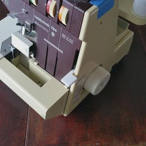 May include: A beige and dark brown serger sewing machine with the text hobbylook 784 electronic and made in West Germany printed on the front. The machine features multiple tension dials, a needle assembly, and a white spool holder on the side.