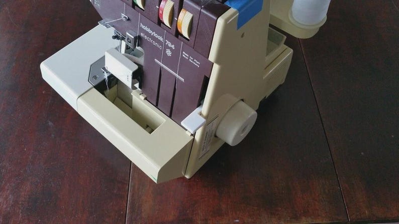 May include: A beige and dark brown serger sewing machine with the text hobbylook 784 electronic and made in West Germany printed on the front. The machine features multiple tension dials, a needle assembly, and a white spool holder on the side.