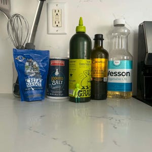 May include: A collection of kitchen items including a blue bag of Celtic Sea Salt, a cylindrical container of Lombardi Iodized Salt, a green squeeze bottle of Grazia Extra Virgin Olive Oil, a bottle of Carapelli Olive Oil, and a bottle of Wesson Vegetable Oil.
