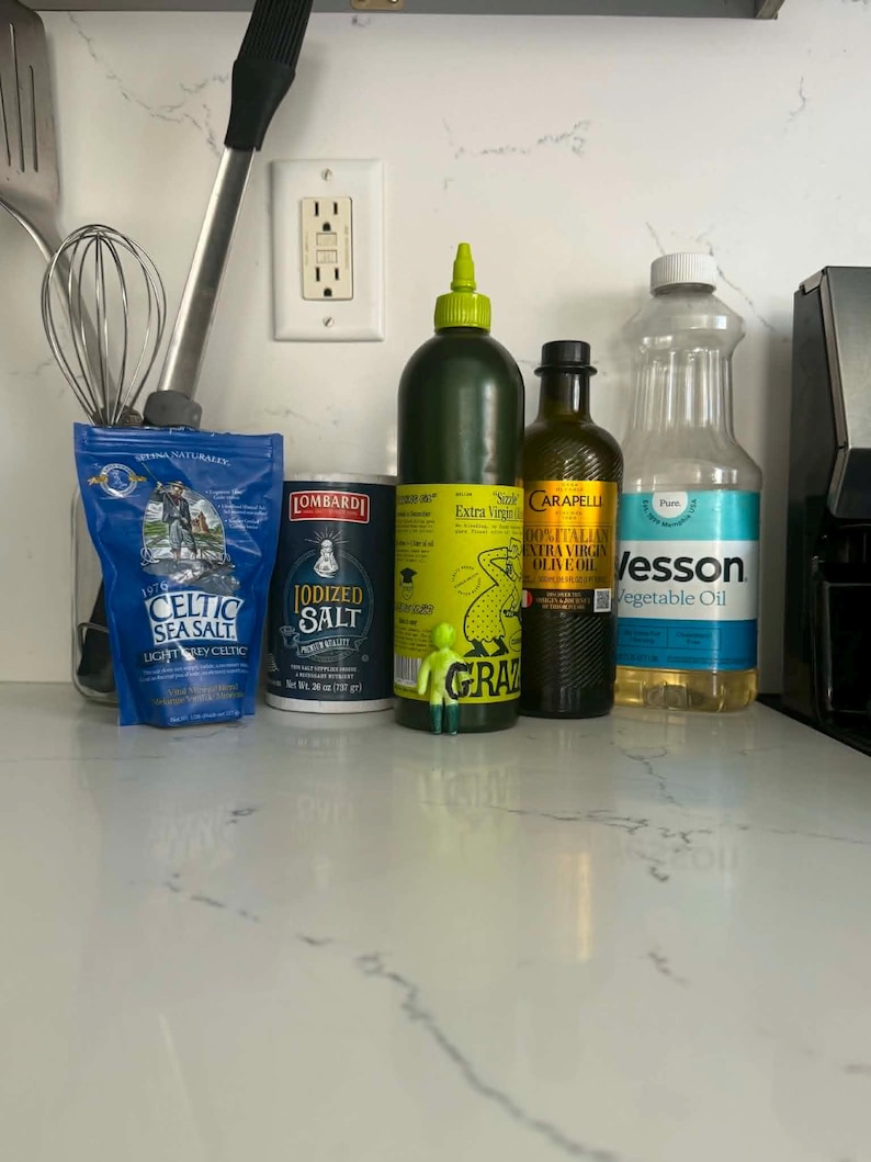 May include: A collection of kitchen items including a blue bag of Celtic Sea Salt, a cylindrical container of Lombardi Iodized Salt, a green squeeze bottle of Grazia Extra Virgin Olive Oil, a bottle of Carapelli Olive Oil, and a bottle of Wesson Vegetable Oil.