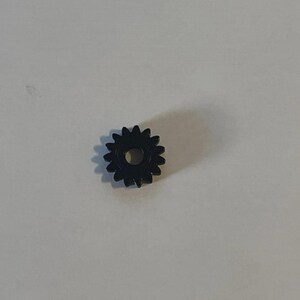 May include: A small, black, circular gear featuring twelve distinct teeth around its outer edge and a centred, circular hole. The gear has a smooth, uniform finish and is positioned flat, displaying its front face and mechanical structure.