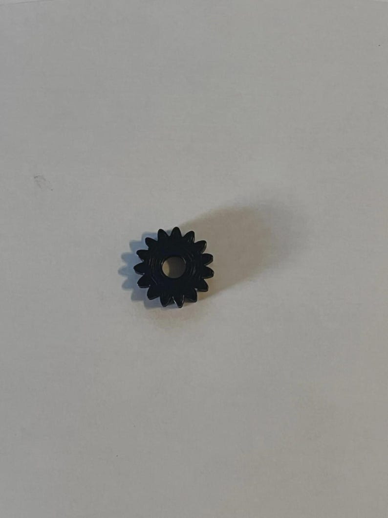 May include: A small, black, circular gear featuring twelve distinct teeth around its outer edge and a centred, circular hole. The gear has a smooth, uniform finish and is positioned flat, displaying its front face and mechanical structure.