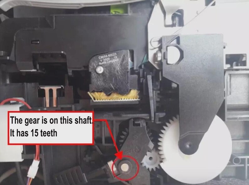 May include: Internal printer mechanism featuring a black plastic housing, a white circular gear, and a yellow brush component. A red arrow points to a gear on a shaft, accompanied by a red-bordered text box stating: The gear is on this shaft. It has 15 teeth.