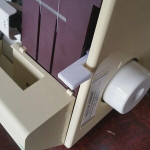 May include: A cream-colored serger sewing machine featuring a dark maroon vertical panel, a white circular adjustment dial on the side, and a white lever. The machine has a visible white thread path and a small rectangular label on the side panel.