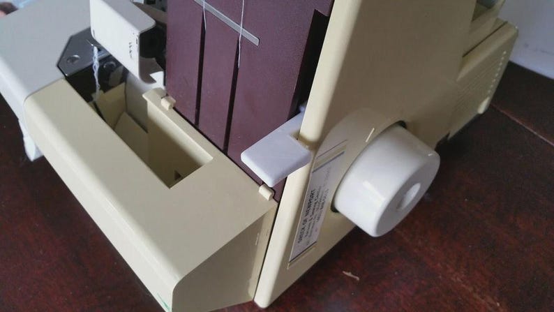 May include: A cream-colored serger sewing machine featuring a dark maroon vertical panel, a white circular adjustment dial on the side, and a white lever. The machine has a visible white thread path and a small rectangular label on the side panel.