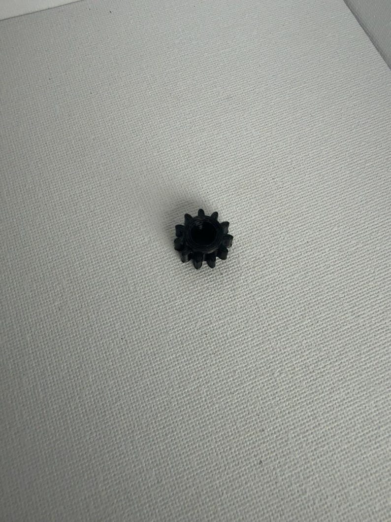 May include: A small, black, circular gear component featuring twelve distinct teeth around its outer perimeter and a hollow, cylindrical center opening, designed for mechanical assembly and rotational movement applications.