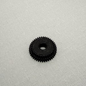 May include: A black, circular, 3D-printed gear featuring a stepped design with two distinct levels of teeth. The component has a smooth, flat top surface with a centered circular hole, showcasing a textured finish from the printing process.