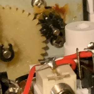 Może przedstawiać: Zbliżenie na biały element elektryczny z srebrnym złączem i czerwonymi i czarnymi przewodami. Element jest przymocowany do płytki drukowanej, a w tle widoczny jest mechanizm zębatkowy.