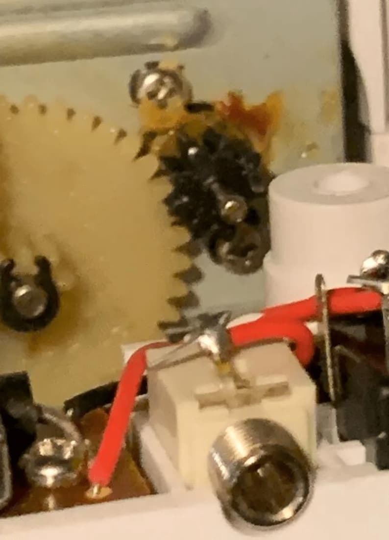 Może przedstawiać: Zbliżenie na biały element elektryczny z srebrnym złączem i czerwonymi i czarnymi przewodami. Element jest przymocowany do płytki drukowanej, a w tle widoczny jest mechanizm zębatkowy.
