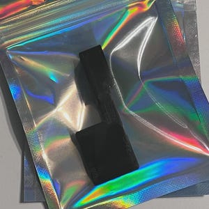 May include: A small, solid black, L-shaped plastic component with a textured surface finish. The object is positioned directly on top of a translucent, iridescent, holographic resealable plastic pouch that reflects vibrant rainbow colors.