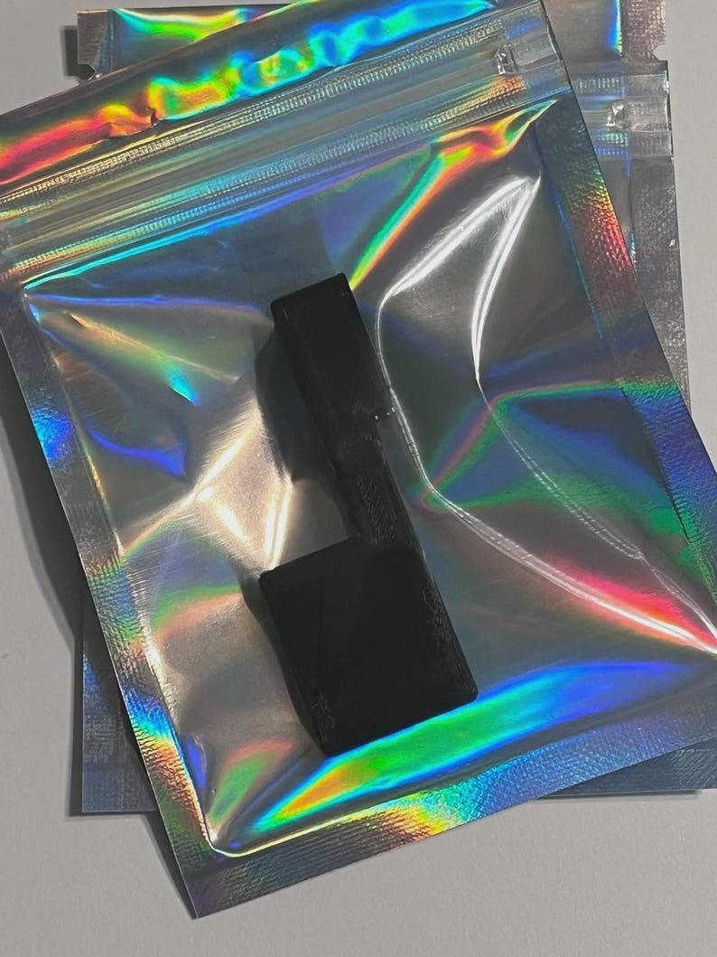 May include: A small, solid black, L-shaped plastic component with a textured surface finish. The object is positioned directly on top of a translucent, iridescent, holographic resealable plastic pouch that reflects vibrant rainbow colors.