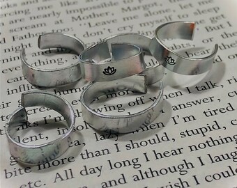 May include: A close-up of several silver rings with engraved designs. The rings are stacked on top of each other and are resting on a book with text. One ring has a lotus flower design.