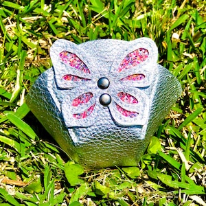 Puede incluir: Brazalete plateado con diseño de mariposa. La mariposa tiene detalles de purpurina rosa y morada y está asegurada con dos pequeños botones grises oscuros. El brazalete está sobre hierba verde.