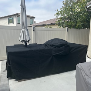May include: A black outdoor furniture cover is covering a large patio table and chairs. The cover is made of a thick, durable material and has a drawstring closure. A gray umbrella is in the background.