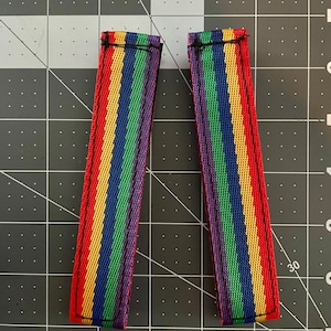 May include: Two rainbow striped fabric straps with black stitching. The straps are approximately 12.7 cm long and 2.5 cm wide.