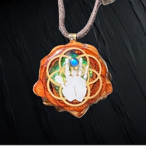 May include: A handcrafted pendant with a unique design. The pendant features a white handprint within a gold-colored geometric pattern, set against a backdrop of orange and green hues. A blue gemstone sits at the top. The pendant hangs from a brown cord.