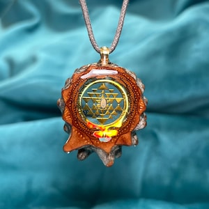 May include: A pendant necklace featuring a gold-colored geometric design encased in a clear resin. The pendant is set within a brown, textured, organic-shaped frame and hangs from a gray cord. The background is a solid teal color.