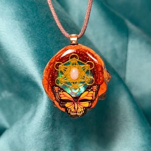 May include: A unique pendant featuring a Metatron's Cube design with a butterfly motif. The pendant has a warm orange and brown base, with gold and iridescent accents. It hangs from a brown cord.