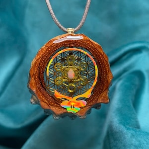 May include: A unique pendant necklace featuring a wood and resin design. The pendant has a gold-colored geometric pattern, a central opal-like stone, and a colorful, stylized butterfly shape. The necklace hangs on a gray cord.